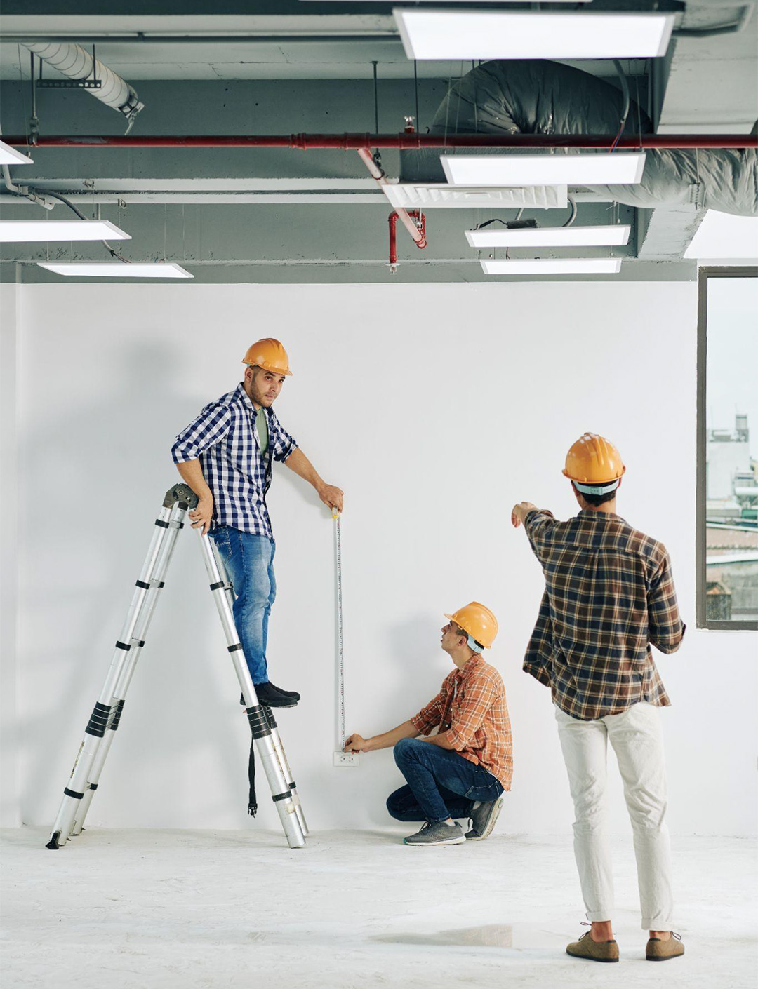 Three construction workers in hard hats measuring a wall in a building under renovation. one stands on a ladder, another holds the tape measure, and the third observes.